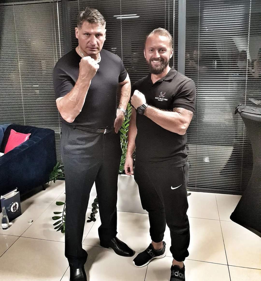 Dwóch mężczyzn w czarnych ubraniach prezentuje napięte bicepsy w nowoczesnym wnętrzu z żaluzjami i niebieską sofą.