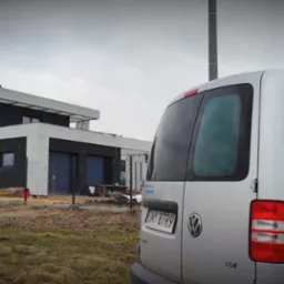 Nowoczesny dom w trakcie budowy, widoczny zza samochodu dostawczego z naklejką reklamową firmy budowlanej Ideal Home Venom, na pierwszym planie ziemia i trawa, w tle pochmurne niebo.