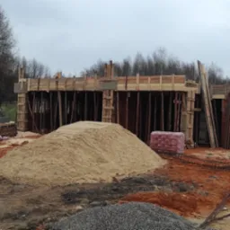 Budowa domu w trakcie realizacji: widoczne fundamenty z drewnianym szalunkiem, sterta piasku, bloczki betonowe i cegły na placu budowy w pochmurny dzień.