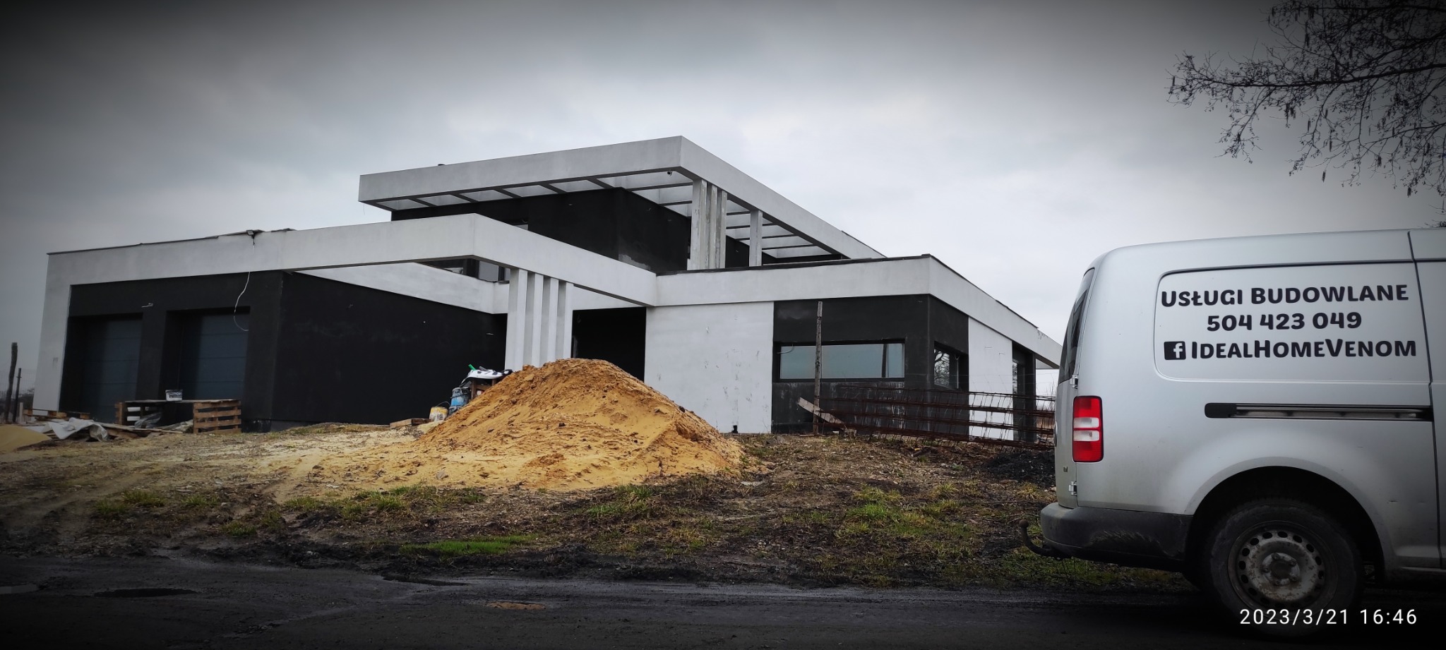 Nowoczesny, częściowo wykończony dom z płaskim dachem w trakcie budowy, z widoczną stertą piasku przed budynkiem i samochodem dostawczym z naklejką reklamową firmy budowlanej na boku.