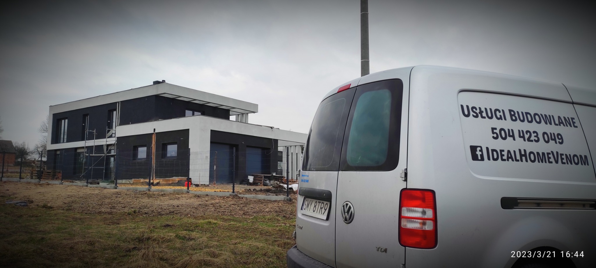 Nowoczesny dom w trakcie budowy, widoczny zza samochodu dostawczego z naklejką reklamową firmy budowlanej Ideal Home Venom, na pierwszym planie ziemia i trawa, w tle pochmurne niebo.