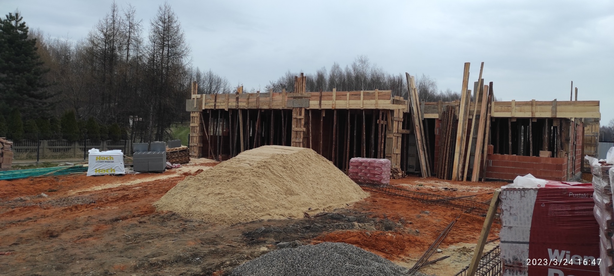 Budowa domu w trakcie realizacji: widoczne fundamenty z drewnianym szalunkiem, sterta piasku, bloczki betonowe i cegły na placu budowy w pochmurny dzień.