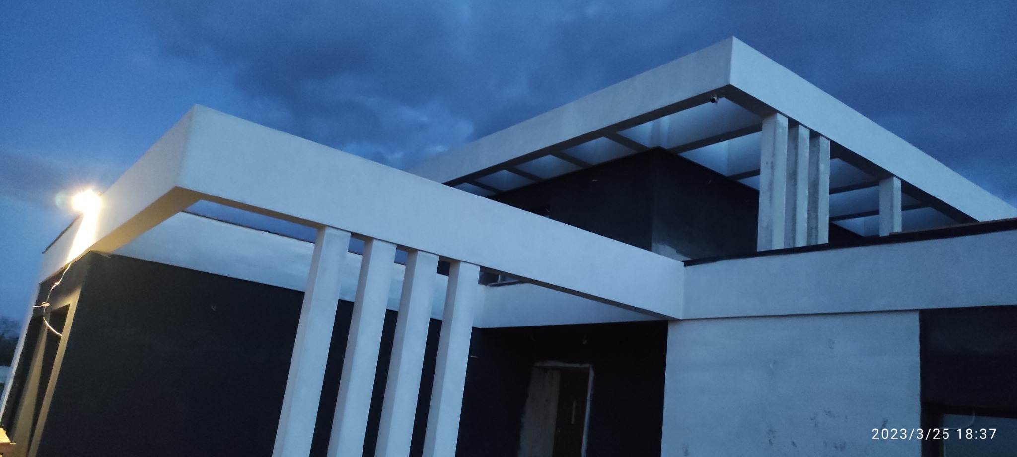 Surowy stan nowoczesnego domu z płaskim dachem i elementami dekoracyjnymi w postaci pionowych belek, sfotografowany o zmierzchu pod pochmurnym niebem.