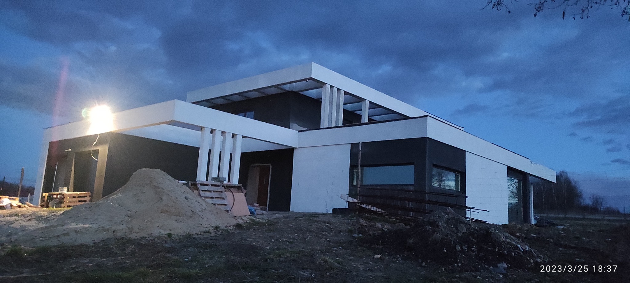 Nowoczesny dom jednorodzinny w trakcie budowy, widoczny pod wieczornym niebem, z hałdą piasku i materiałami budowlanymi na pierwszym planie, oświetlony reflektorem.