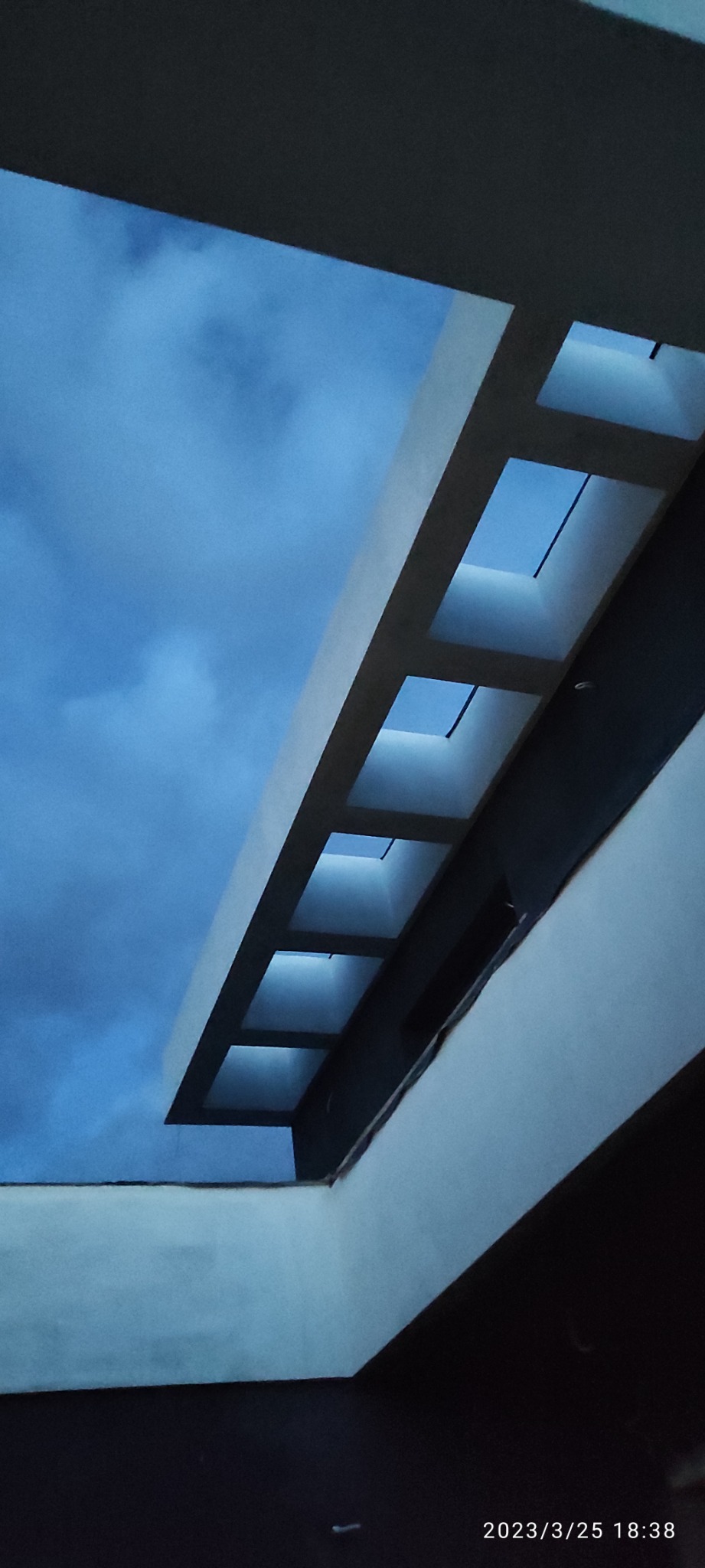 Wnętrze domu w trakcie budowy, widok z dołu na strop z otworami okiennymi, przez które widać pochmurne, błękitne niebo.