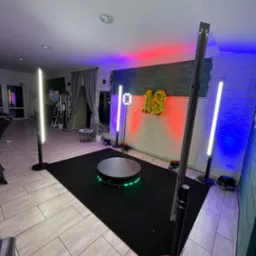 Okrągła platforma obrotowa z zielonymi diodami LED na czarnym dywanie, otoczona przez lampy podłogowe z białym i kolorowym światłem, złoty balon w kształcie liczby 18 na tle ceglanej ściany.