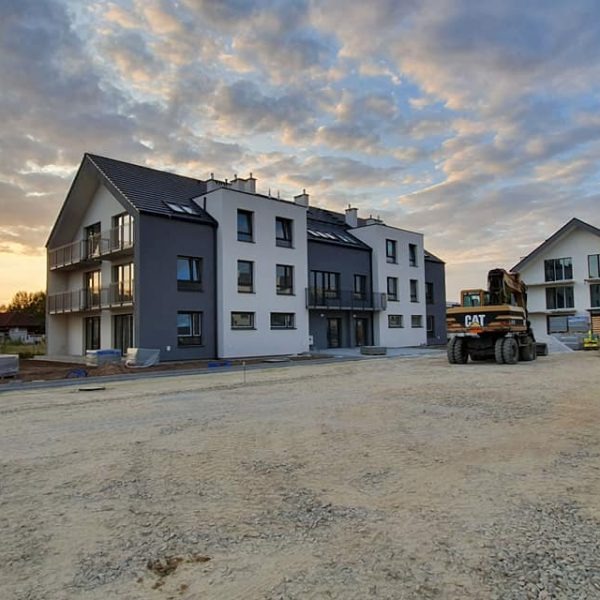 Nowoczesny, trzypiętrowy dom murowany z balkonami, szarą elewacją i ciemnym dachem, widziany z perspektywy placu budowy z koparką CAT w tle, pod błękitnym, zachmurzonym niebem.