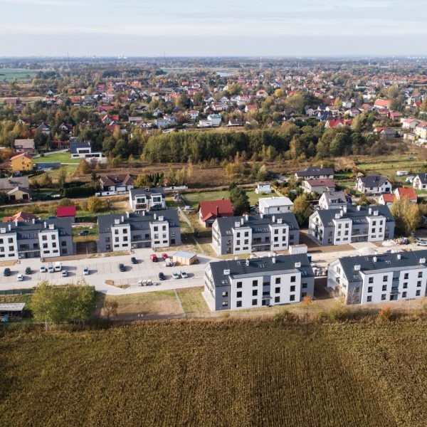 Nowoczesne, dwukondygnacyjne domy murowane z szarymi dachami i białymi elewacjami w Oławie, widziane z lotu ptaka, z parkingiem przed budynkami i polami w tle.