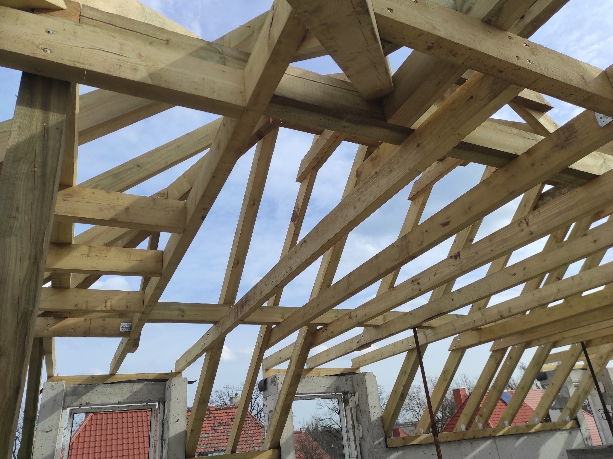 Surowa więźba dachowa z jasnego drewna, widok od dołu na tle błękitnego nieba z chmurami, fragment muru z widocznym oknem oraz dachy okolicznych budynków w tle.