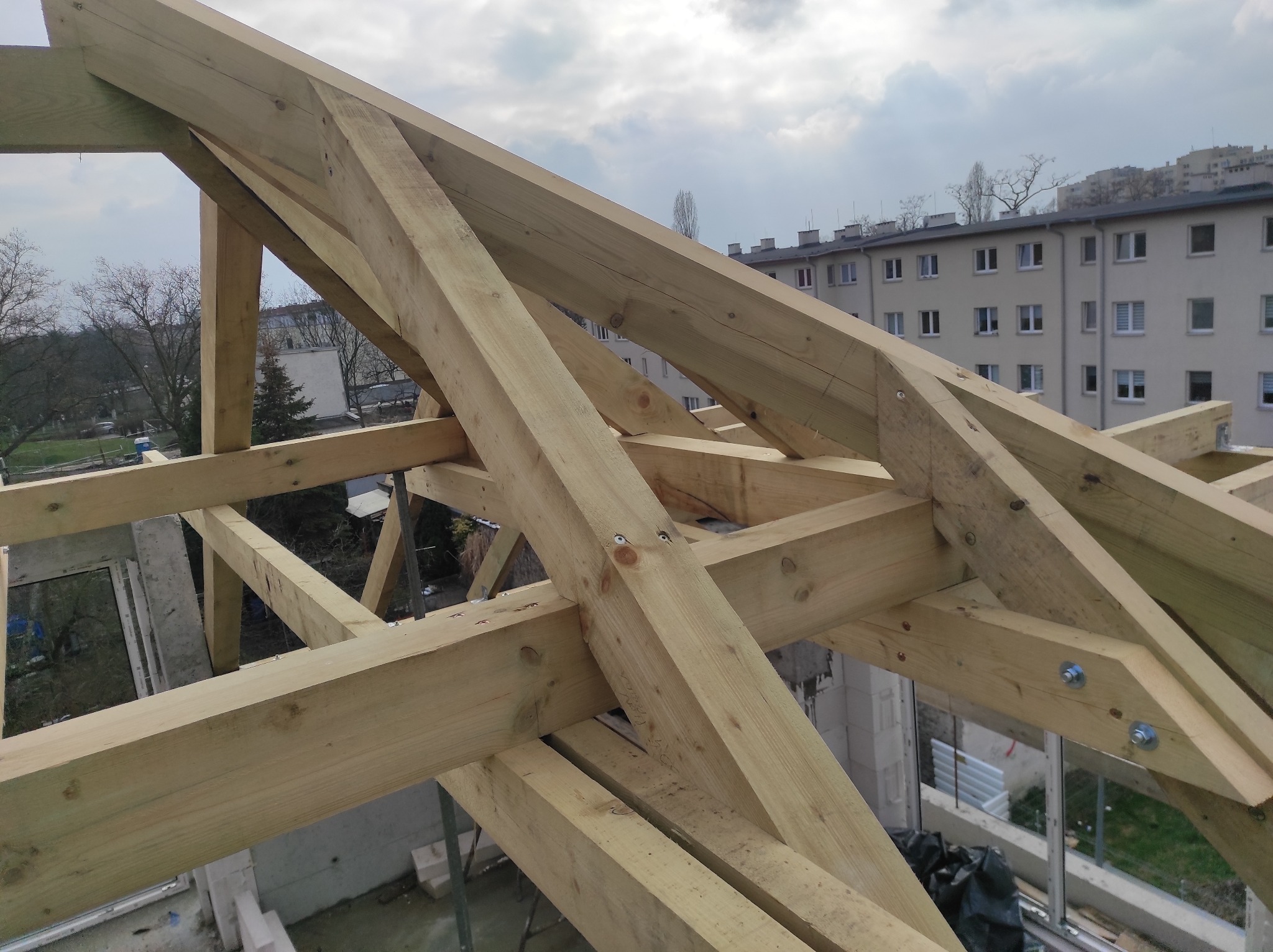 Surowa, drewniana konstrukcja więźby dachowej z widocznymi elementami mocującymi, na tle budynku mieszkalnego i pochmurnego nieba.
