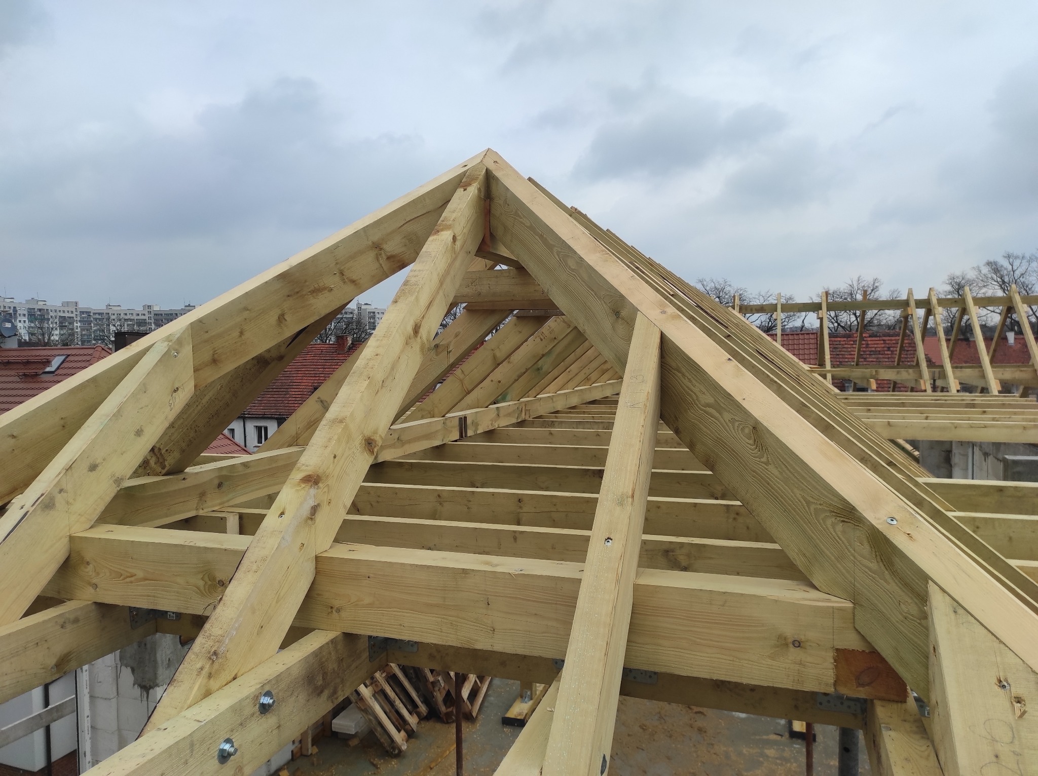 Surowa drewniana konstrukcja więźby dachowej z widocznymi śrubami i metalowymi łącznikami, na tle częściowo widocznych budynków mieszkalnych i pochmurnego nieba.