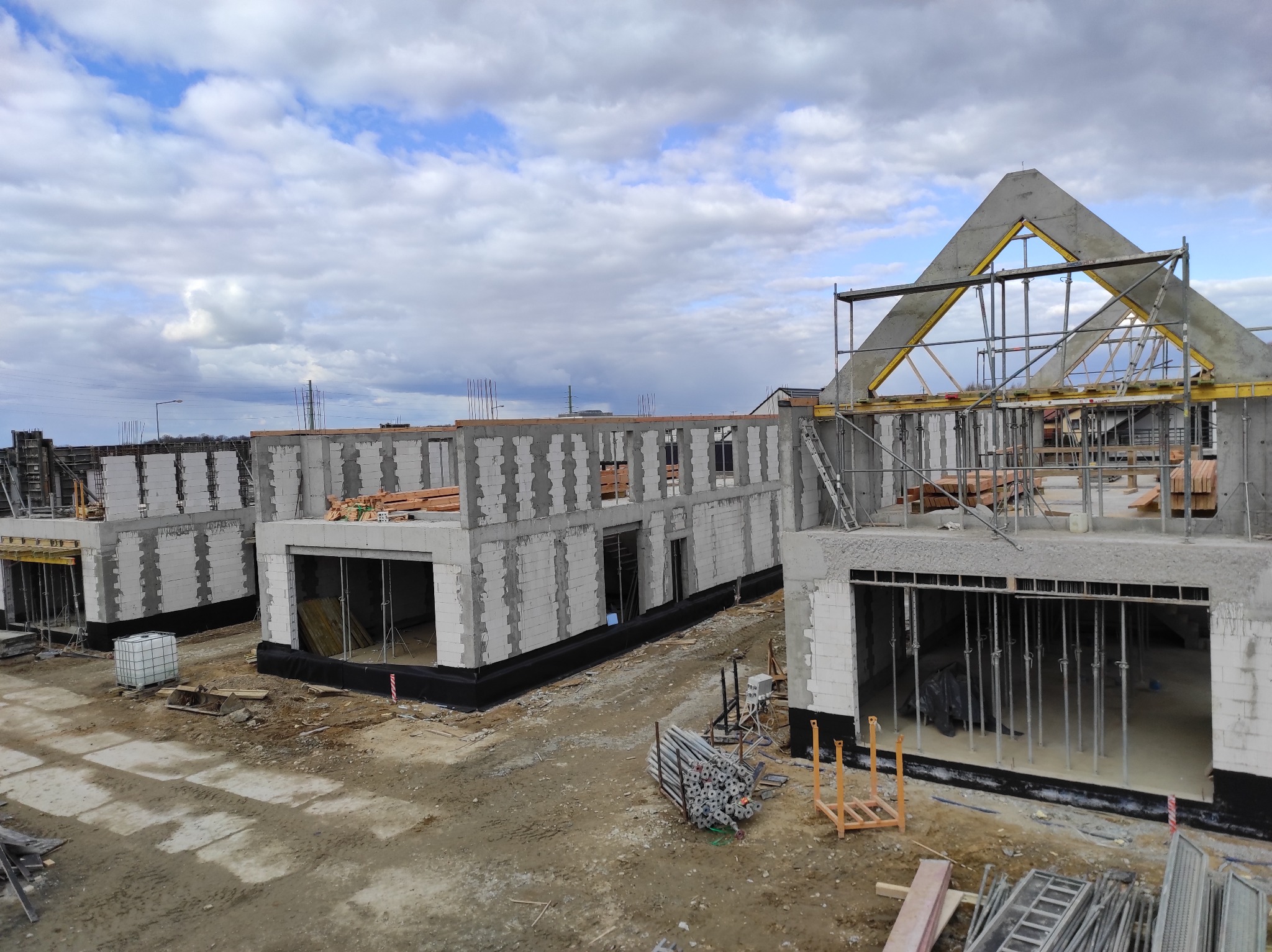Budowa kilku domów murowanych z bloczków betonu komórkowego w stanie surowym otwartym, widoczne rusztowania i elementy konstrukcji dachu, pochmurne niebo w tle.