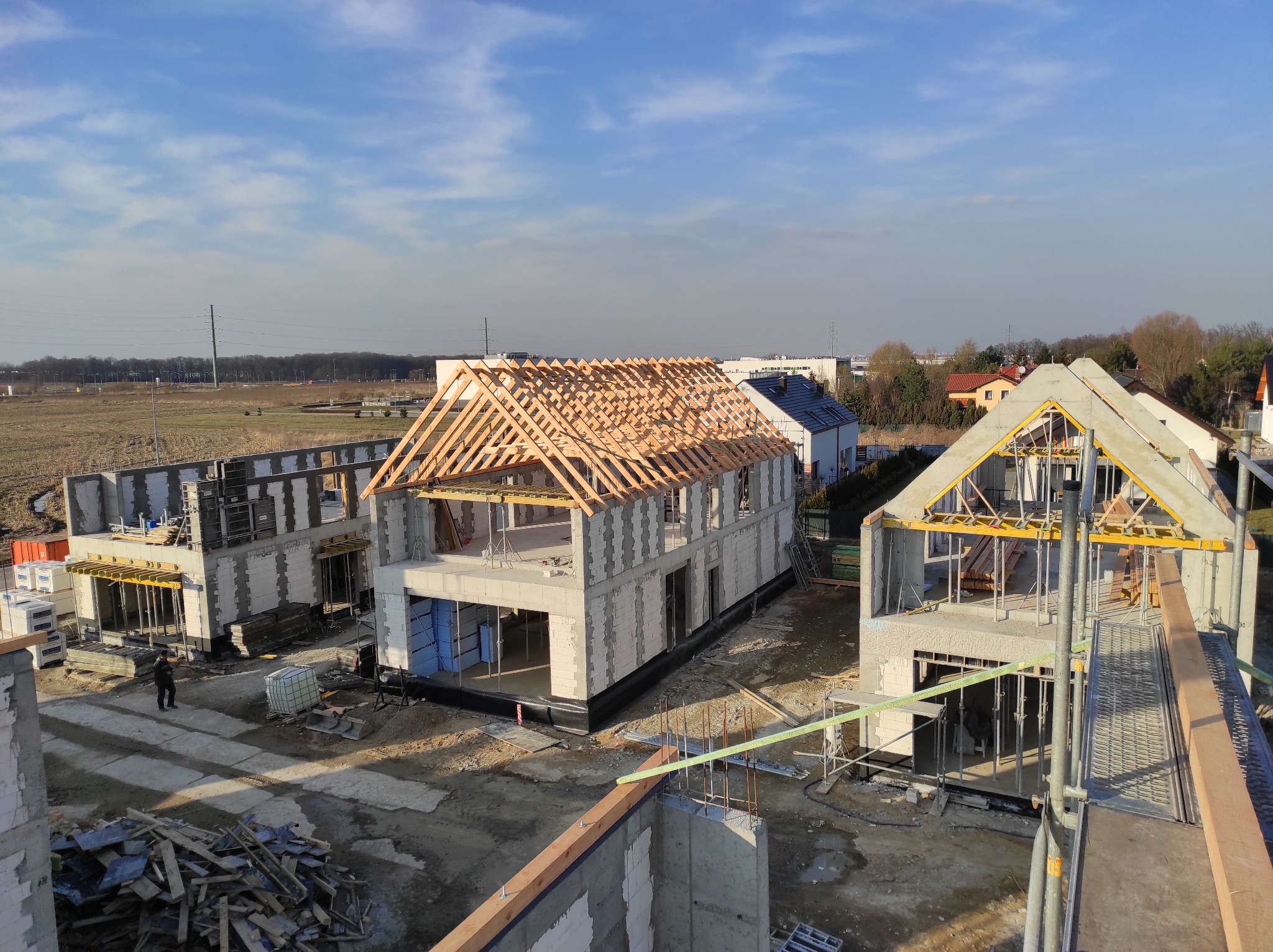 Trzy domy w trakcie budowy z widoczną więźbą dachową, wykonane z białych bloczków, na jednym z nich widać już czarne pokrycie dachu, widok z góry na plac budowy w słoneczny dzień.