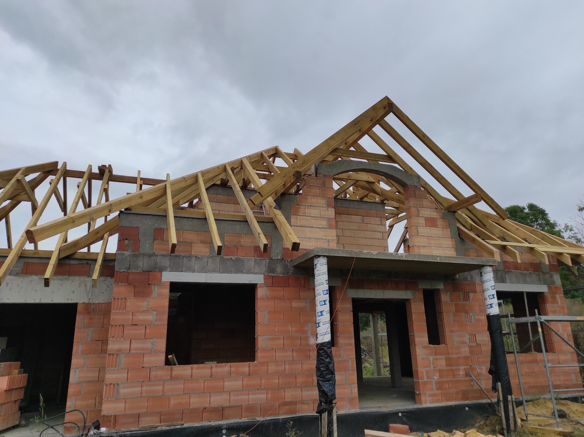 Surowy stan domu murowanego z czerwonej cegły, z widoczną drewnianą konstrukcją dachu i elementami betonowymi, pod pochmurnym niebem.
