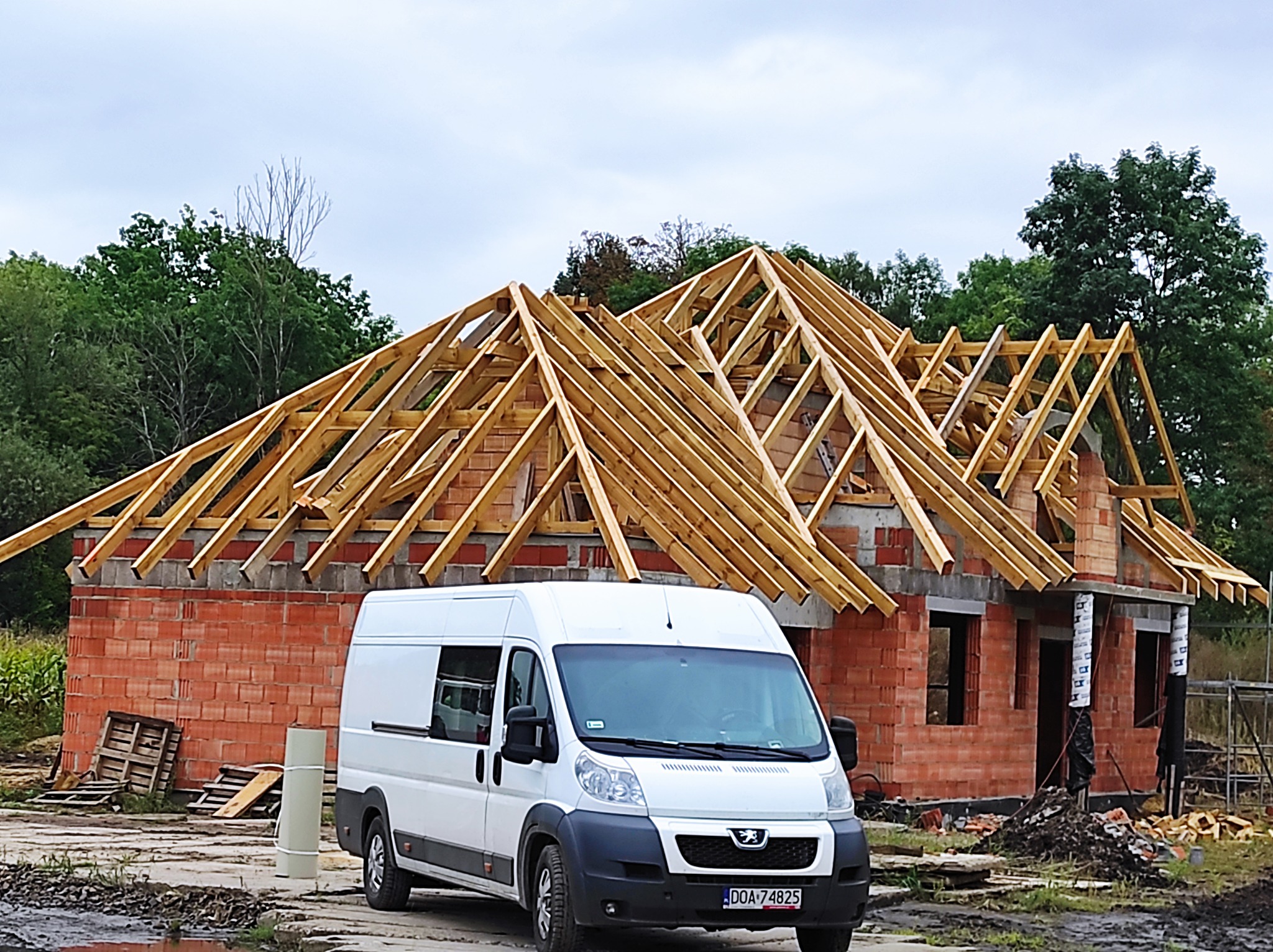 Surowy stan domu murowanego z czerwoną cegłą i drewnianą konstrukcją dachu w trakcie budowy, z białym samochodem dostawczym zaparkowanym przed budynkiem.