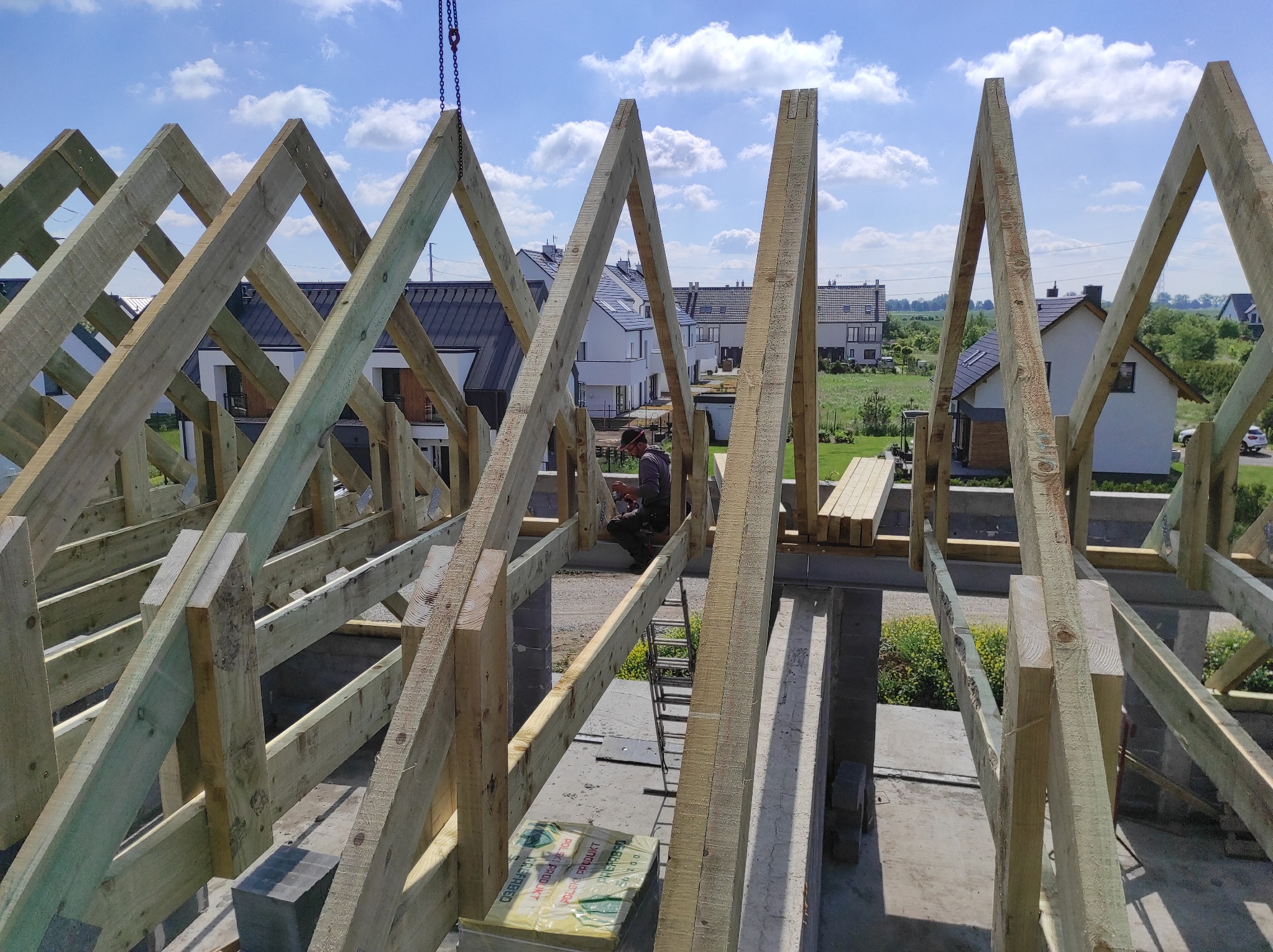 Konstrukcja drewniana więźby dachowej w trakcie montażu, widoczne elementy nośne i pracownik na tle budynków mieszkalnych.