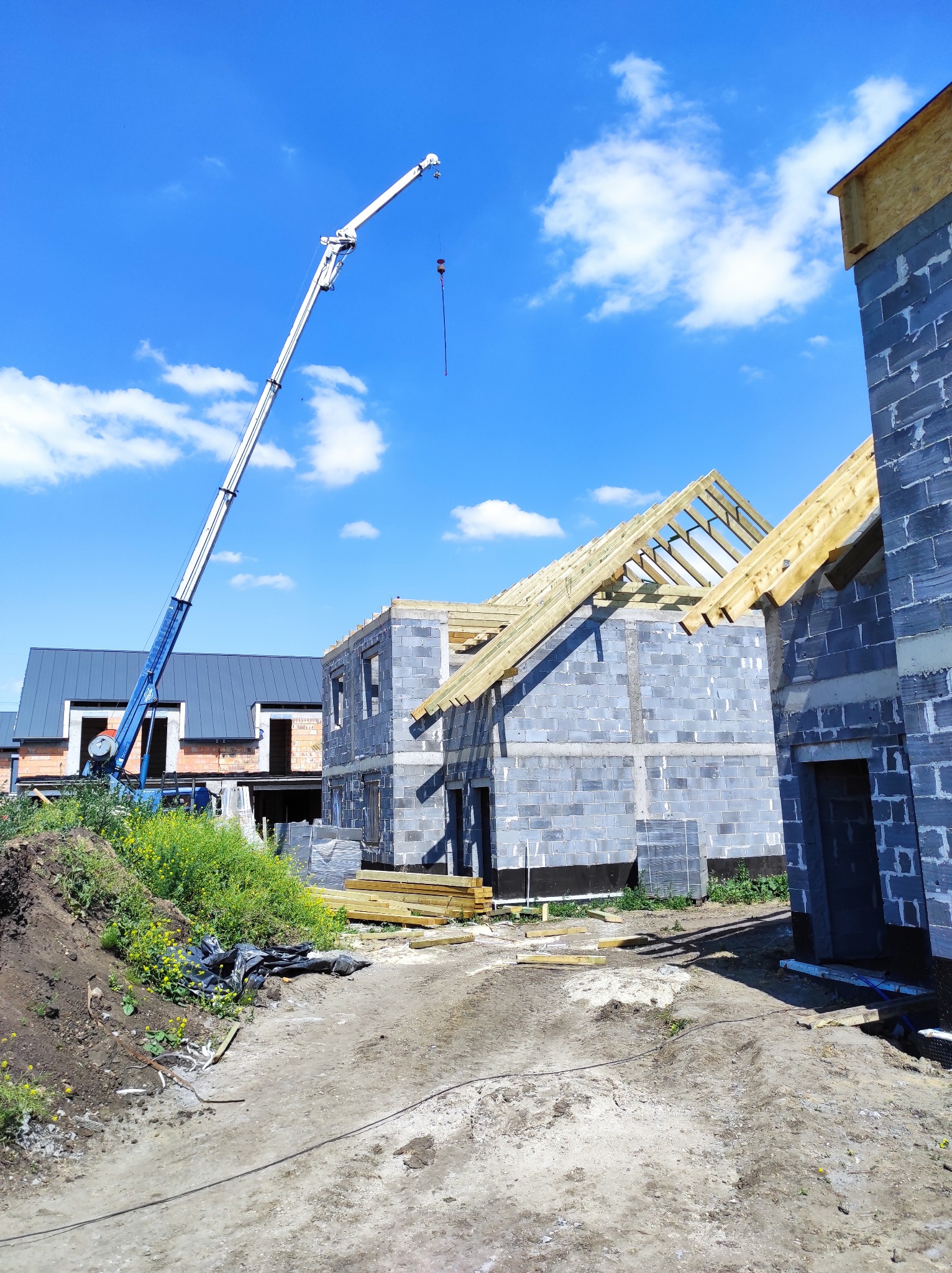 Budowa domu z bloczków, widoczna konstrukcja dachu z drewnianych belek, na placu budowy stoi dźwig, w tle inne budynki z szarą blachą na dachu, niebieskie niebo z chmurami.