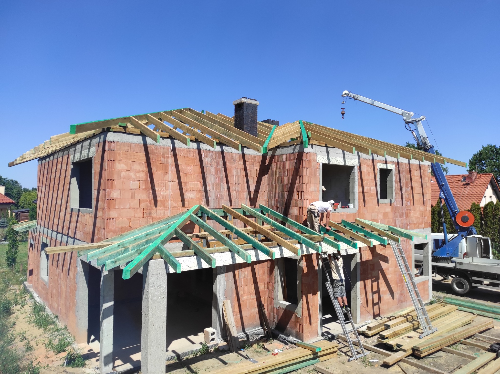 Budowa domu murowanego z czerwonej cegły: montaż drewnianej więźby dachowej z zieloną impregnacją, widoczny dźwig i pracownicy na drabinach.