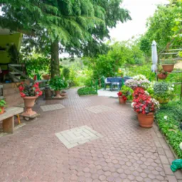 Urokliwy ogród z brukowanym patio, rabatami kwiatowymi w donicach, małym oczkiem wodnym i zielonymi krzewami, tworzący przytulną przestrzeń rekreacyjną przy domu z zieloną elewacją.