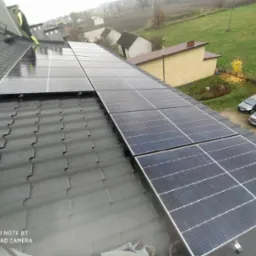 Montaż paneli słonecznych na dachu z ciemnej dach&oacute;wki, widoczny pracownik w odblaskowej kurtce, tło z zabudową jednorodzinną i jesiennym krajobrazem.