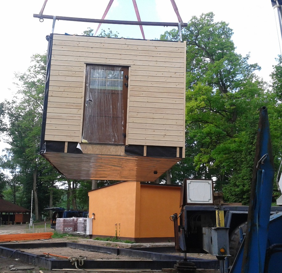 Moduł domu z drewnianą elewacją, z drzwiami zabezpieczonymi folią, podnoszony dźwigiem nad przygotowanym fundamentem i pomarańczowym budynkiem.