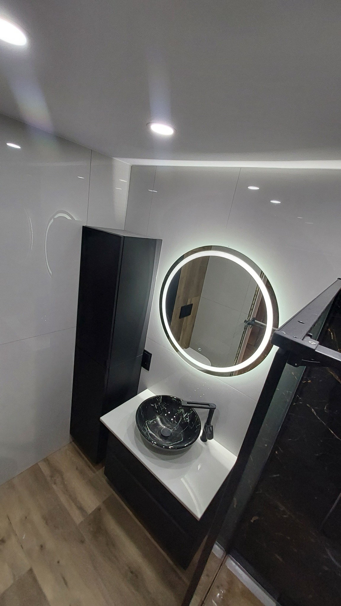 Nowoczesna łazienka z czarną szafką, blatem i umywalką w marmurkowy wzór, okrągłe lustro z podświetleniem LED i białe płytki na ścianach.