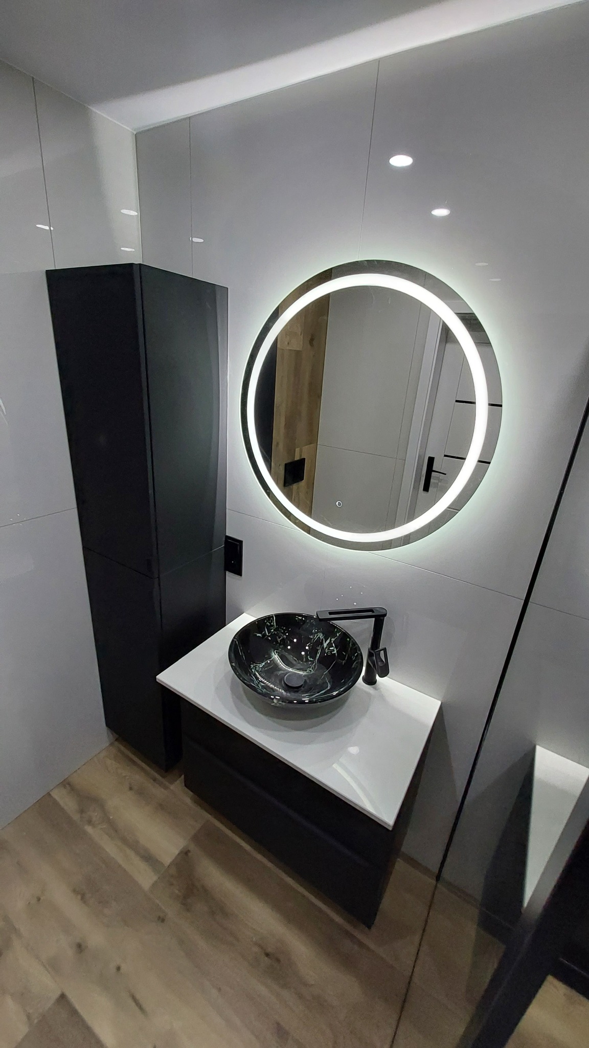 Nowoczesna łazienka z czarną, marmurkową umywalką nablatową, podświetlanym okrągłym lustrem, białą szafką z czarnymi frontami oraz wysoką, czarną szafką boczną.