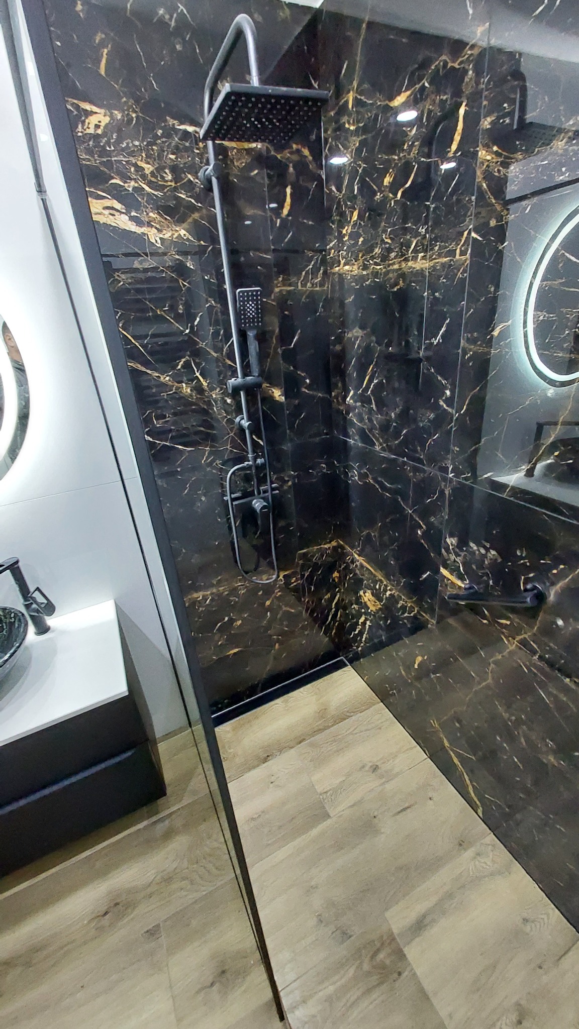 Nowoczesna kabina prysznicowa z czarnymi płytkami imitującymi marmur ze złotymi żyłami, drewnopodobną podłogą i czarną armaturą.