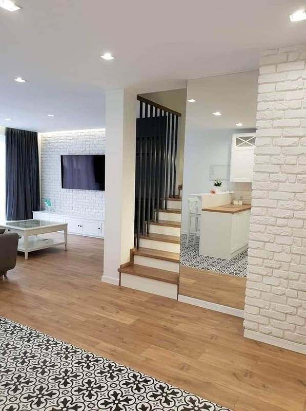 Jasne, przestronne wnętrze domu z drewnianą podłogą, schodami z czarną balustradą i ścianą z białej cegły, z lustrem wbudowanym w ścianę odbijającym fragment kuchni.