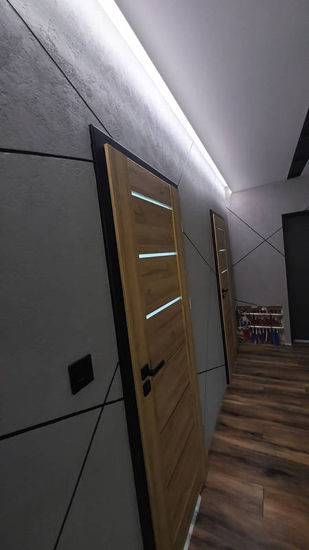Szara ściana z dekoracyjną fakturą i czarnymi liniami, drewniane drzwi z trzema poziomymi, podświetlanymi paskami, oświetlenie LED podsufitowe, podłoga z paneli imitujących drewno.