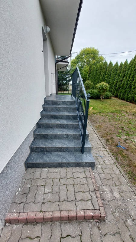 Zewnętrzne schody wejściowe wyłożone płytkami imitującymi kamień, z metalową balustradą, prowadzące do drzwi wejściowych budynku z elewacją w jasnym kolorze.