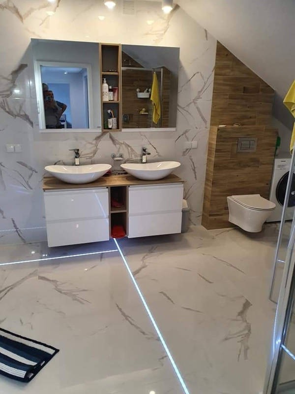 Nowoczesna łazienka na poddaszu z dwoma owalnymi umywalkami nablatowymi, podświetlaną podłogą LED, marmurowymi płytkami ściennymi i drewnianą zabudową toalety.