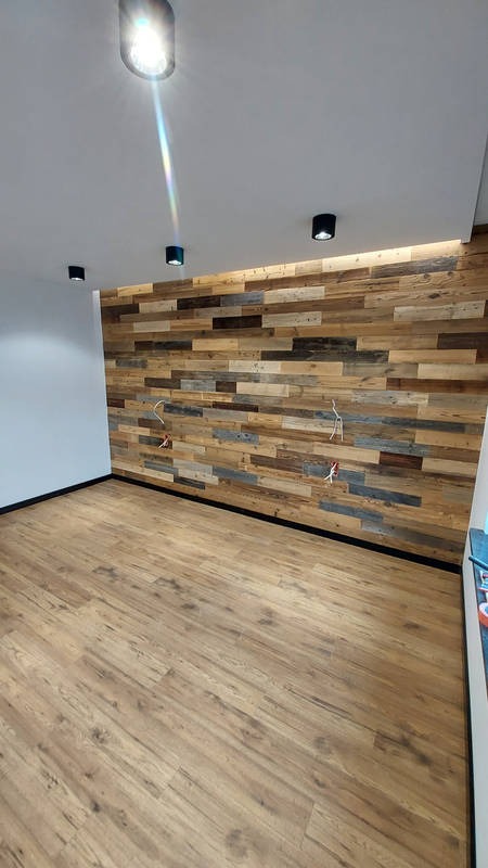 Pokój z drewnianą ścianą z desek o różnej kolorystyce, laminowaną podłogą, białymi ścianami i czarnymi reflektorami sufitowymi; widoczne wystające przewody elektryczne.