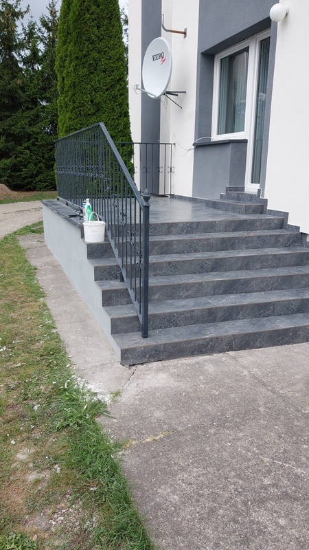 Wykończone, grafitowe schody wejściowe z metalową barierką, na których stoi białe wiadro z narzędziami i preparatami, widoczna antena satelitarna na elewacji budynku.