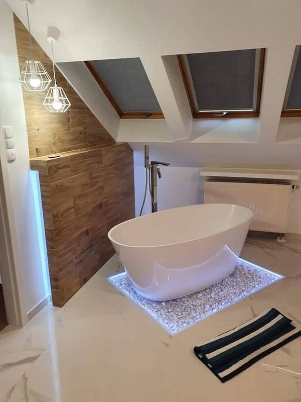 Nowoczesna łazienka na poddaszu z wolnostojącą białą wanną owalną, umieszczoną na podświetlanym podeście z białych kamyków. W tle widoczne okna dachowe i drewniana ściana.