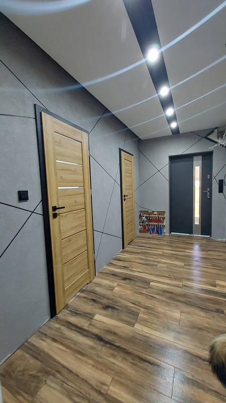 Nowoczesny przedpokój z drewnianą podłogą imitującą deski, szarą ścianą z geometrycznym wzorem, dwiema drewnianymi drzwiami z czarnymi ościeżnicami oraz sufitem z oświetleniem punktowym.