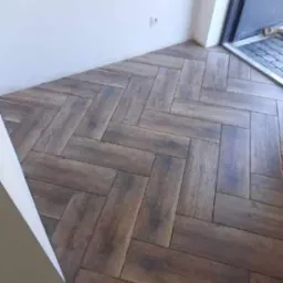 Ułożona w jodełkę, ciemna, drewnopodobna glazura na podłodze w rogu pomieszczenia z białymi ścianami i drzwiami balkonowymi z szarą wycieraczką.