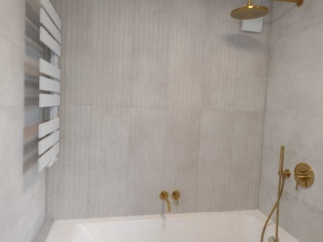 Wykończona łazienka z wanną, grzejnikiem drabinkowym, deszczownicą i złotą armaturą na tle szarych, pionowo ułożonych płytek.