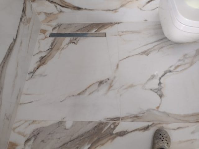 Ułożona podłoga z płytek imitujących marmur w łazience, widoczny odpływ liniowy i fragment toalety, w kadrze fragment stopy w gumowym obuwiu.