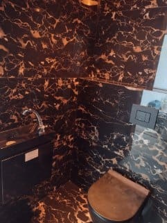 Ciemna łazienka wykończona czarnym marmurem z brązowymi żyłkami, widoczna umywalka z szafką, bateria i toaleta. Na ścianie przyciski do spłukiwania.