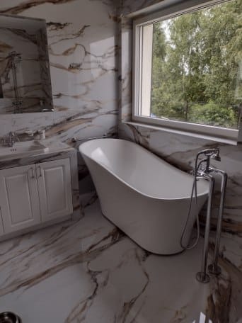 Nowoczesna łazienka z wolnostojącą białą wanną na tle marmurowych płytek podłogowych i ściennych z brązowymi żyłami, widok na zewnątrz przez okno.