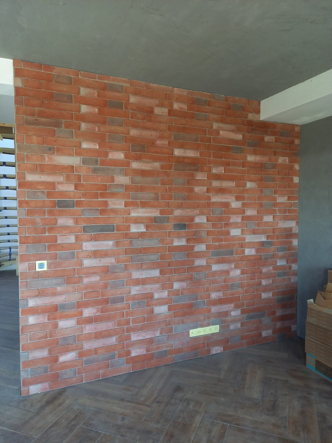 Ściana z cegieł klinkierowych w odcieniach czerwieni i szarości, ułożona na podłodze w jodełkę, z widocznym fragmentem szarego sufitu i elementami instalacji elektrycznej.