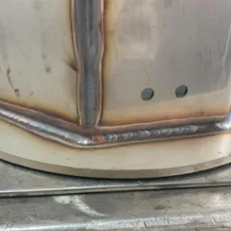 Szczegółowe zbliżenie spoiny na metalowym zbiorniku z widocznymi otworami, wykonanej metodą spawania łukowego, z charakterystycznymi przebarwieniami termicznymi.