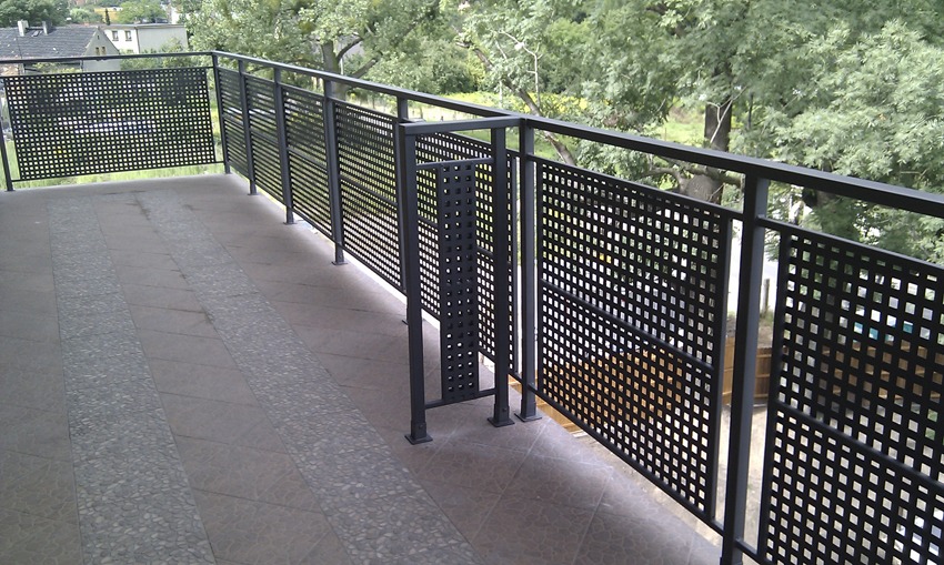 Nowoczesna, metalowa balustrada balkonowa z kwadratowymi otworami, szara płytka podłogowa z dekoracyjnym pasem z kamyczków, widok na zieleń drzew w tle, minimalizm w designie.