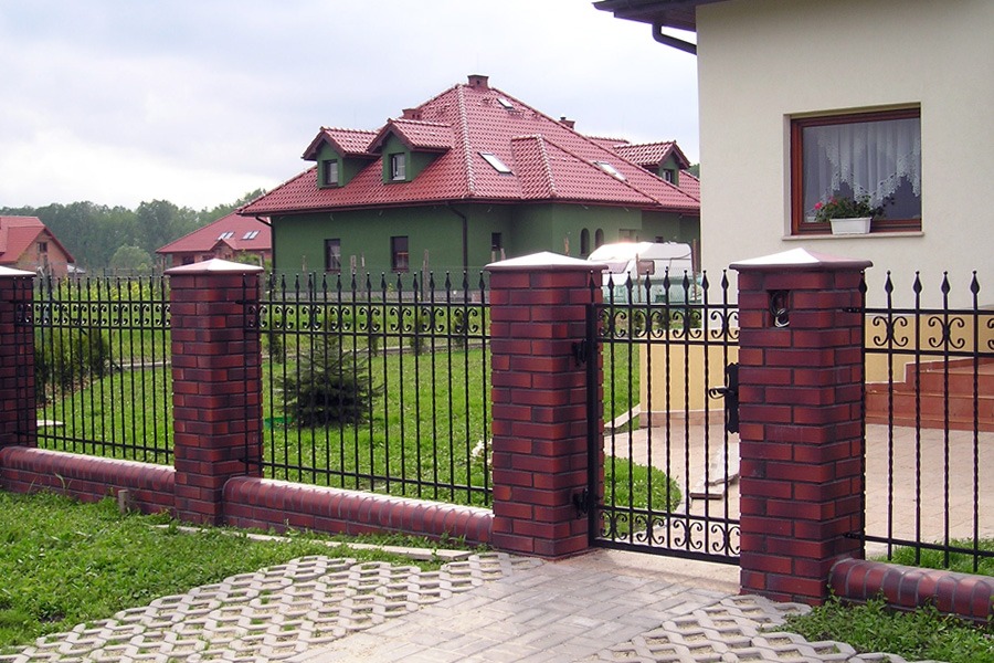 Czarna, metalowa brama i ogrodzenie z dekoracyjnymi elementami, osadzone na ceglanych słupkach i podmurówce, na tle zielonego trawnika i zabudowy jednorodzinnej.