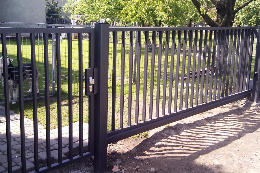 Nowoczesna, przesuwna brama metalowa z pionowymi prętami, zamek z gałką, widoczny fragment ogrodzenia i trawnika w tle, cień bramy na ziemi.