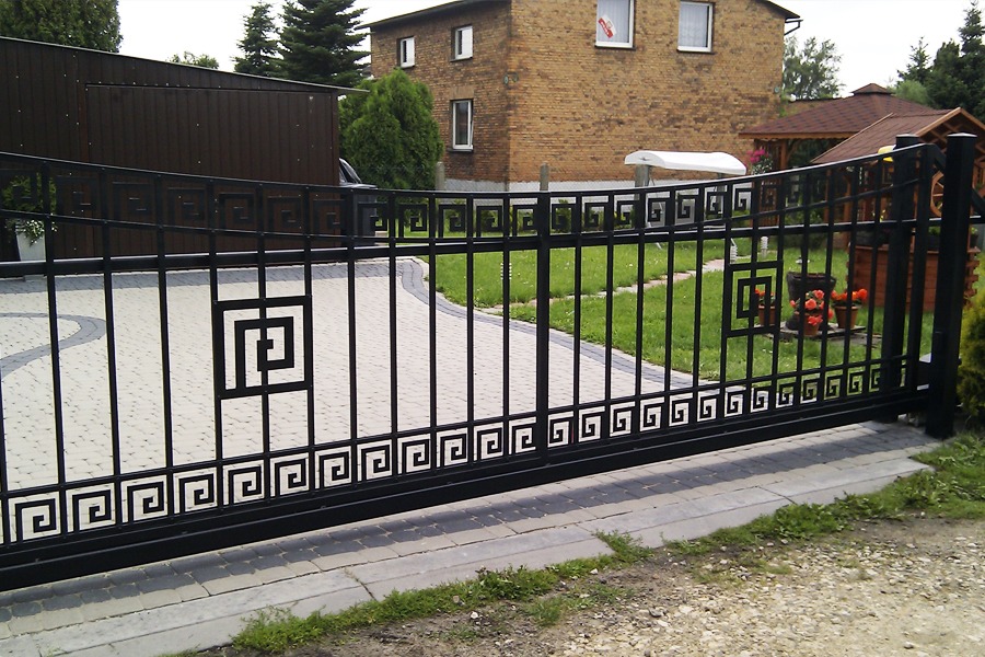 Czarna, przesuwna brama metalowa z geometrycznym wzorem greckim i kwadratowymi zdobieniami, na tle brukowanej podjazdów i zielonego trawnika z elementami architektury ogrodowej