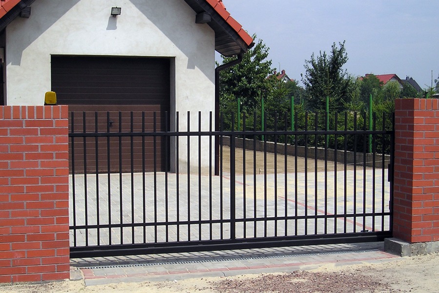 Przesuwna brama metalowa w kolorze czarnym, zamontowana między dwoma słupkami z czerwonej cegły, na tle białego garażu z brązową bramą segmentową.