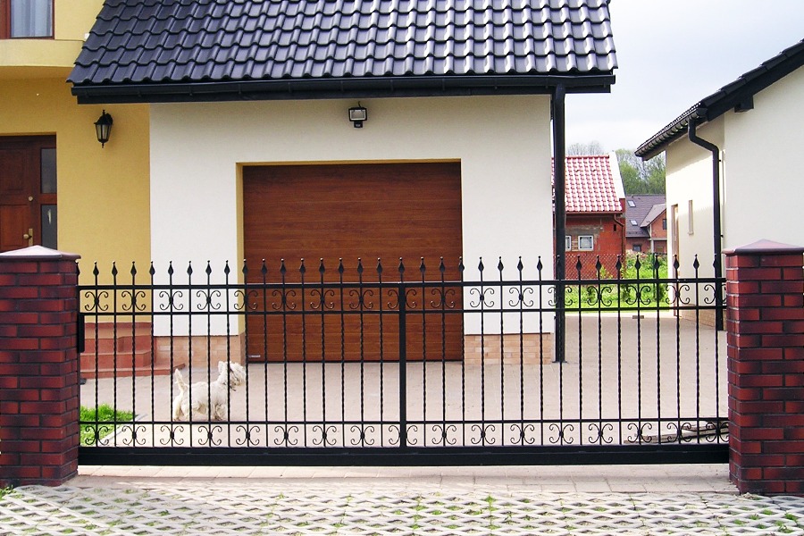 Czarna, metalowa brama przesuwna z dekoracyjnymi elementami, osadzona pomiędzy dwoma słupkami z czerwonej cegły, na tle domu z brązową bramą garażową i mały biały pies za ogrodzeniem.