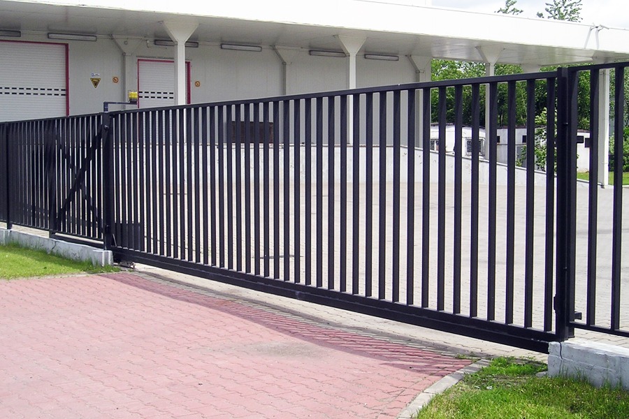 Czarna, przesuwna brama metalowa z pionowymi prętami, zamontowana przed budynkiem magazynowym z bramami rolowanymi i kostką brukową.